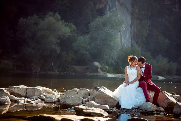 Retrato sensível do noivo em terno vermelho beijando a noiva adorável no ombro enquanto sentado na pedra perto do rio e da floresta . — Fotografia de Stock