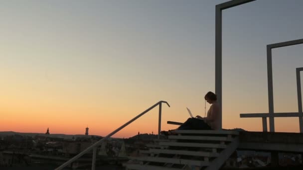 Silhueta de jovem mulher de negócios elegante segurando laptop ao pôr do sol na ponte da cidade. Paisagem urbana fundo — Vídeo de Stock