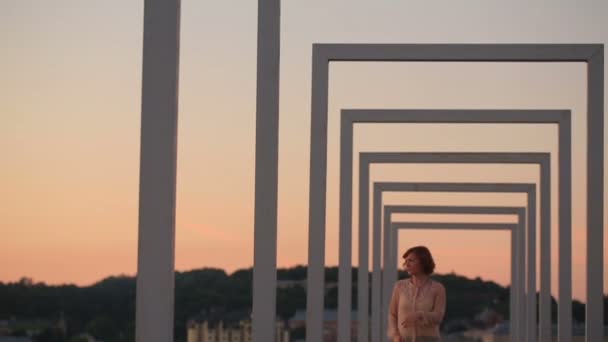 Linda mujer de moda urbana joven caminando en el puente de la ciudad de alta tecnología en la puesta del sol — Vídeo de stock