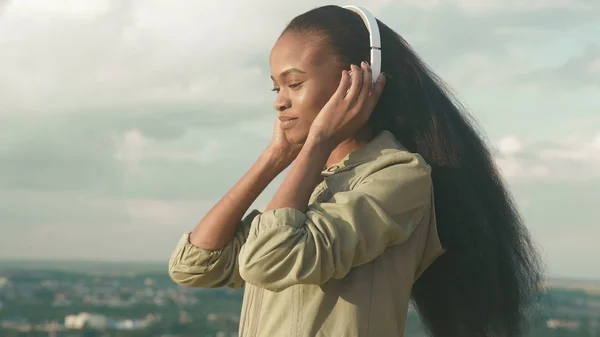 Linda menina afro-americana ouvir música e gosta. Sorrindo jovem menina negra no fundo da cidade turva — Fotografia de Stock