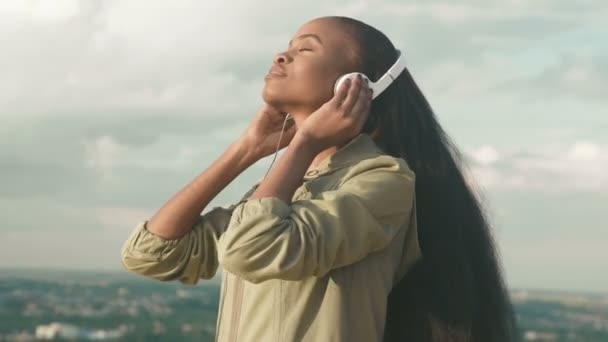 Африканская девочка слушает музыку и получает удовольствие. Улыбающаяся молодая черная девушка на фоне размытого города — стоковое видео