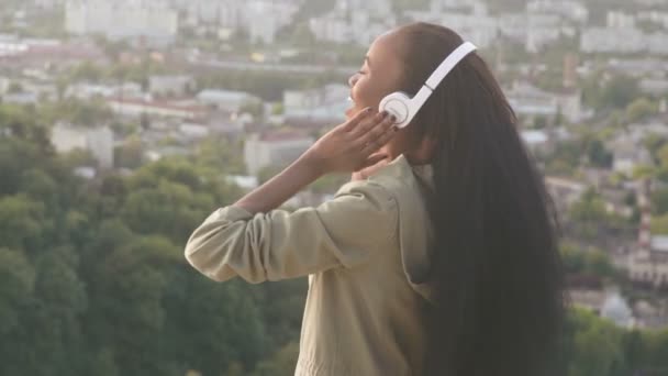 Widok z tyłu piękne african american Girl słuchać muzyki i cieszy. Uśmiechnięta młoda czarna dziewczyna na tle miasta niewyraźne — Wideo stockowe