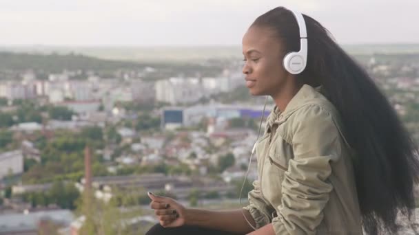 Mooie jonge Afro-Amerikaanse meisje luisteren naar muziek en glimlachen. Mooi zwart meisje buiten op cityscape achtergrond — Stockvideo