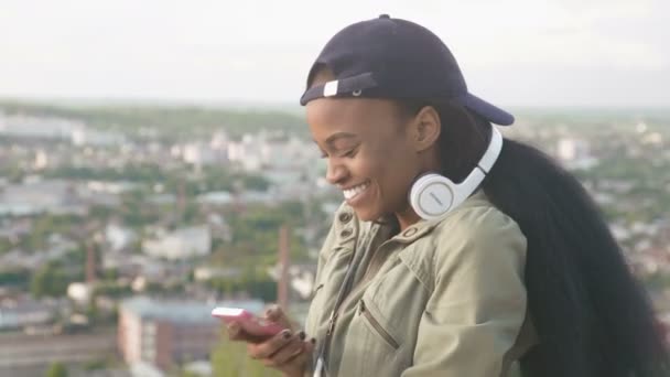 Χαριτωμένο αφρικανική αμερικανική κορίτσι στο καπάκι ακούει μουσική στα ακουστικά και να χαμογελά. Cityscape θολή φόντο — Αρχείο Βίντεο
