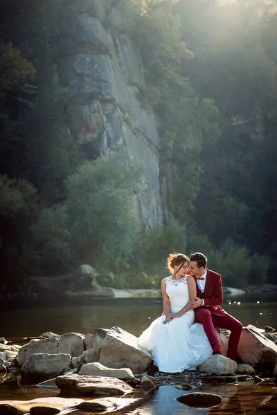 Retrato vertical de la sensible pareja de recién casados abrazados tocando las narices y descansando sobre la piedra cerca del río . — Foto de Stock