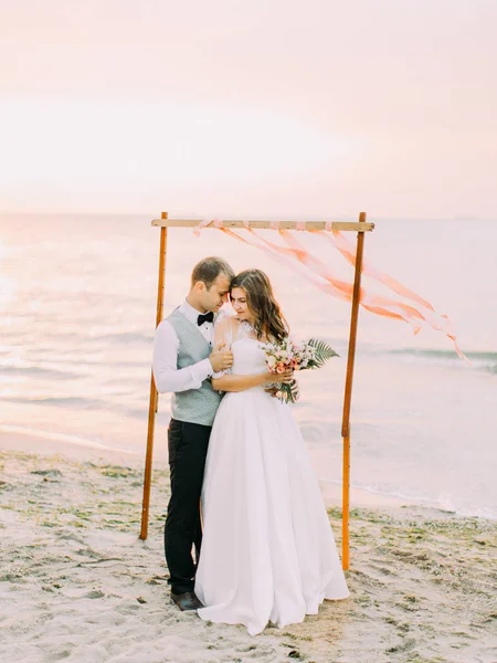 La vista completa del novio feliz abrazando a la novia cerca del arco de la boda en la playa. La composición del atardecer . — Foto de Stock