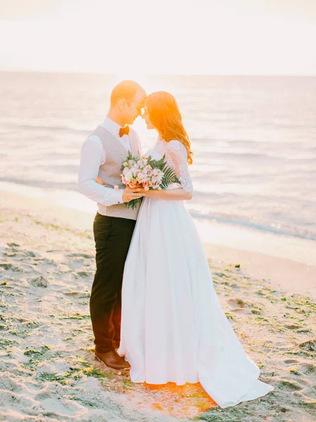 在日落时分, 可爱的新婚夫妇站在大海的背景上, 长着头对头的全景. — 图库照片