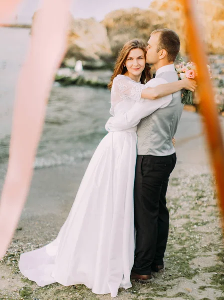 Вид молодоженов в полный рост. Жених целует невесту в голову на фоне моря . — стоковое фото