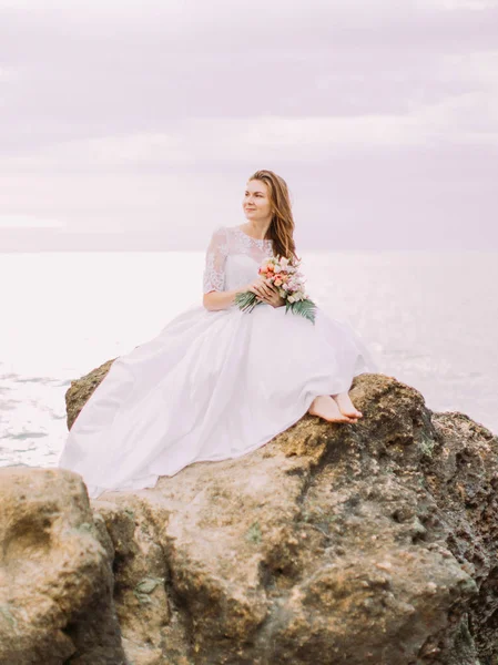 La sposa in abito da sposa lungo è seduto sulla scogliera e guardando il tramonto sullo sfondo del mare . — Foto Stock