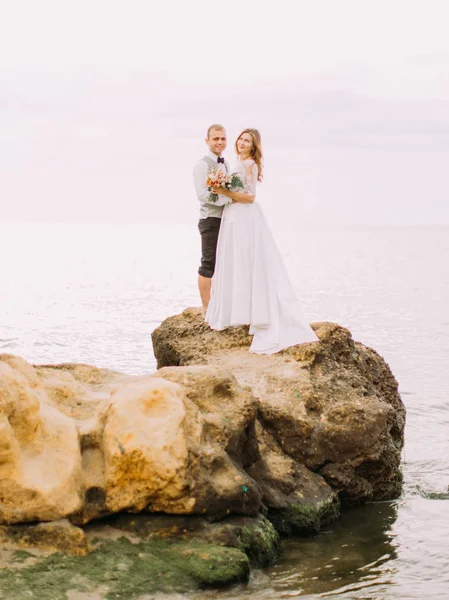 Precioso retrato al aire libre en la longitud completa de los recién casados de pie en el acantilado en el fondo del mar . — Foto de Stock