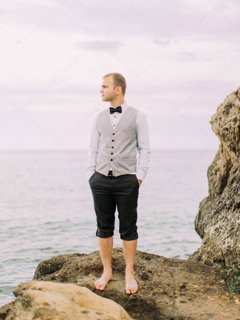 The stylishly dressed groom in the wedding suit is keeping his hands in the pockets and looking at the right side while standing on the cliff at the background of the sea.