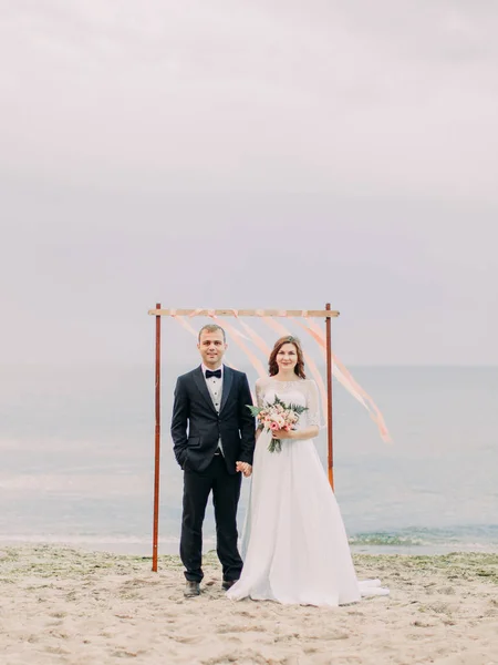 Vista completa de los recién casados tomados de la mano en el fondo de los palos con cintas rosas en la playa . — Foto de Stock