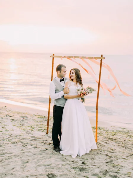 El novio sonriente está abrazando a la novia cerca del arco de la boda colocado en la playa durante la puesta del sol . — Foto de Stock