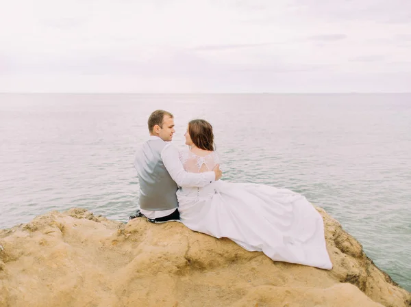 Romantikus Hátulnézet a csak házas ül a szikla között a tenger alatt a naplementében. — Stock Fotó