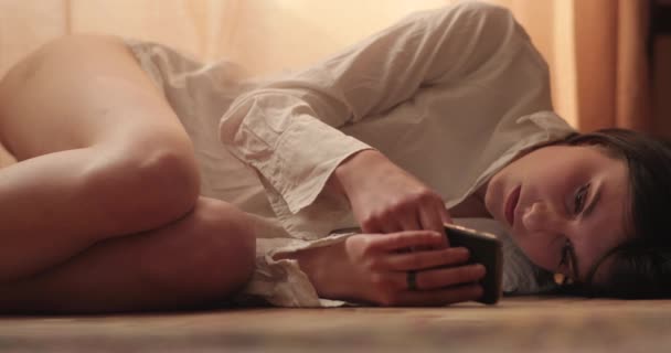 Uma mulher bonita está conversando em redes sociais em seu telefone, deitada em um cobertor em um chão de madeira, em um apartamento no sótão. 4k tiro . — Vídeo de Stock