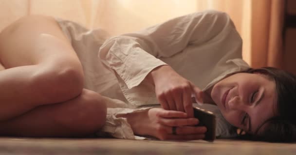 Een mooie glimlachende vrouw kletst in sociale netwerken op haar telefoon, liggend op een deken op een houten vloer, in een loft appartement. 4k Schot. — Stockvideo