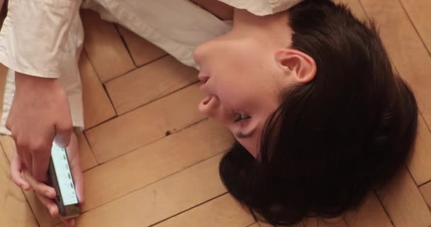 Een mooie brunette is aan het chatten in sociale netwerken op haar telefoon, liggend op een deken op een houten vloer, in een loft appartement. 4k Schot. — Stockvideo