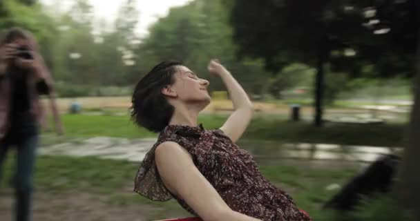 Video van gelukkige aantrekkelijke vrouw draaiend op een schommel in de regen. Lachend meisje met kort brunette haar heeft plezier in de speeltuin terwijl de regen valt. 4k video. — Stockvideo