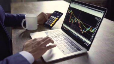 Masanın üzerinde bir dizüstü bilgisayar var. Masada oturan ve dizüstü bilgisayarda çalışan bir iş adamı. Laptop ekranında istatistik ve grafikler var. Akıllı telefondan hesap makinesiyle iş adamını kapat.