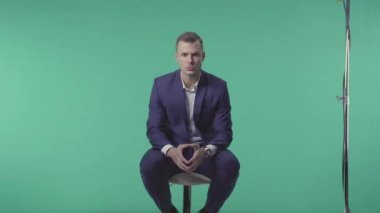 Mavi takım elbiseli güçlü yüzlü genç sakallı işadamı yeşil arka planda fotoğrafçıya poz veriyor. Taburede oturuyor ve kameraya bakıyor. 4k 'da sahne arkasında..