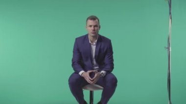 Mavi takım elbiseli güçlü yüzlü genç sakallı işadamı yeşil arka planda fotoğrafçıya poz veriyor. Taburede oturuyor ve kameraya bakıyor. 4k 'da sahne arkasında..