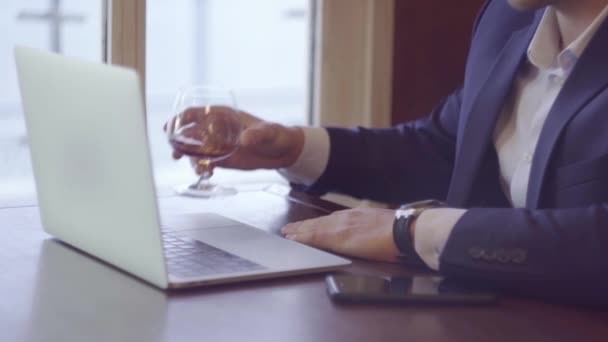 Modny biznesmen w niebieskim garniturze, siedzący przed oknem i pracujący na laptopie obok stołu stoi szklanka whisky. Praca w domu. — Wideo stockowe
