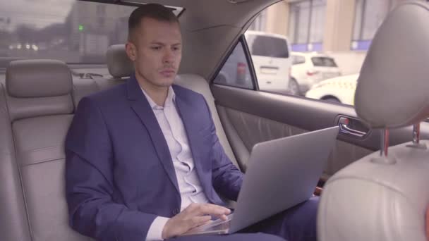 Mavi takım elbiseli bir iş adamı hareket halindeki arabanın arka koltuğunda oturuyor ve ofise giderken dizüstü bilgisayarla çalışıyor. Yakışıklı bir erkek, güçlü bir yüzü ve sarı saçları var.. — Stok video