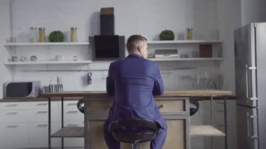 İşadamı şık bir mutfakta mavi takım elbisesiyle oturuyor, kafasını pencereye çeviriyor ve bir şeyler düşünüyor. Yakışıklı sarışın adam evde çalışıyor. Şık ve modern mutfak..
