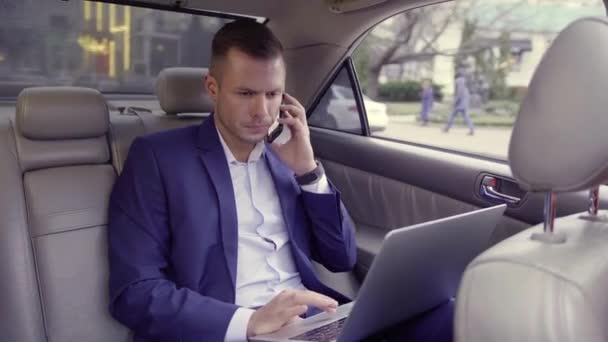 Portret obchodníka v modrém obleku sedí na zadním sedadle jedoucího auta, telefonuje, zatímco pracuje na notebooku, zatímco míří do kanceláře. Hezký vousatý pes se silnou tváří a blond vlasy v — Stock video