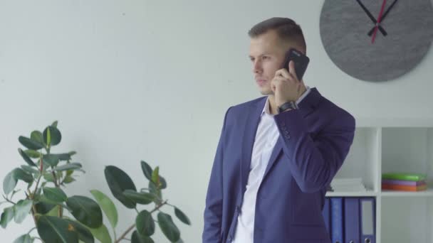 Feliz hombre de negocios en traje azul hablando por teléfono móvil en la oficina con estilo. Guapo rubia chico hablando teléfono emocionalmente en remoto lugar de trabajo . — Vídeo de stock