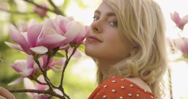 Портрет молодой очаровательной женщины с великолепными золотыми волосами среди цветочного дерева с розовыми лепестками. Привлекательная юная леди расслабляется на природе. 4K видео . — стоковое видео