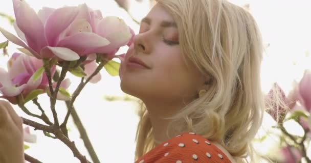 ピンクの花びらを持つ花の木の間で壮大な黄金の髪を持つ若い魅力的な女性の肖像画。魅力的な若い女性は、自然の中でゾッとしている。4kビデオ. — ストック動画