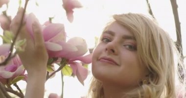 Pembe yapraklı çiçek ağacının arasında, muhteşem altın sarısı saçları olan çekici bir kadının yakın portresi. Güzel sarışın kız çiçek açan ağacın yanında poz veriyor, gülümsüyor ve sevimli çiçekleri kokluyor. 4k