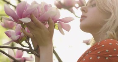 Pembe yapraklı çiçek ağacının arasında, muhteşem altın sarısı saçları olan çekici bir kadının yakın portresi. Güzel sarışın kız çiçek açan ağacın yanında poz veriyor, inanılmaz çiçeklere hayran ve onları kokluyor.