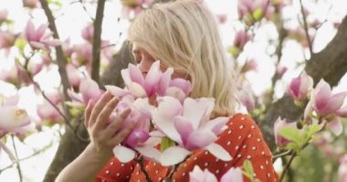 Pembe yapraklı çiçek ağacının arasında, muhteşem altın saçlı, çekici bir kadının portresi. Çiçek açan ağacın yanında poz veren güzel sarışın kız inanılmaz çiçeklere hayran ve onları kokluyor. 4k