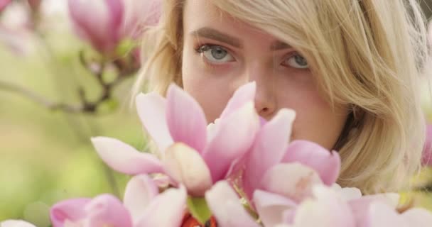 Nära porträtt av ung charmig kvinna med ett magnifikt gyllene hår, bland blomsterträd med rosa kronblad. Härlig blond flicka poserar nära blommande träd, beundrar otroliga blommor och sniffa dem — Stockvideo