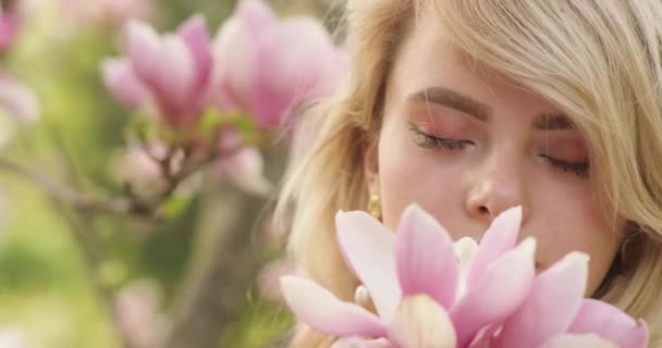 Close portrait of young charming woman with a magnificent golden hair, among flower tree with pink petals. Lovely blonde girl posing near blooming tree, admiring incredible flowers and sniffing them — Stock Video