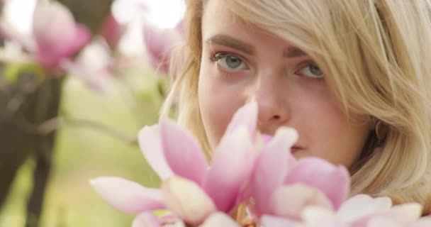 Blízký portrét mladé okouzlující ženy s nádhernými zlatými vlasy, mezi květinami s růžovými okvětními lístky. Krásná blondýnka pózuje blízko kvetoucího stromu, usmívá se a čichá k rozkošným květinám. 4k — Stock video