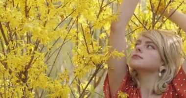 Sarı çiçeklerin arasında poz veren çekici sarışın kızın narin portresi. Muhteşem altın sarısı saçları olan çekici bir kadın doğada serinliyor. 4k video.