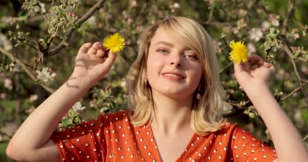 Ritratto ravvicinato di una giovane donna sorridente con i capelli corti biondi che guarda dritto davanti alla macchina fotografica, in piedi vicino all'albero in fiore in una giornata di sole. Il suo viso è incredibilmente bello con un sorriso affascinante e blu — Video Stock