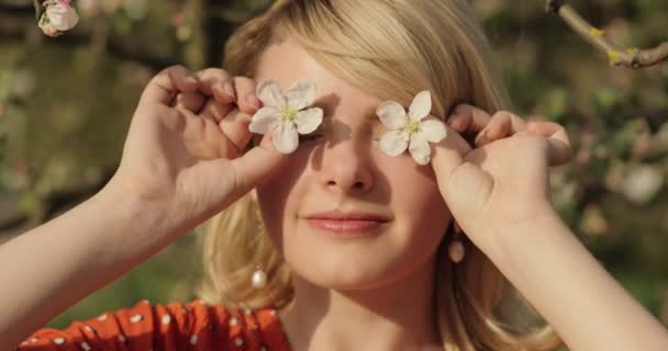 Detailní portrét mladé usměvavé ženy s krátkými blond vlasy dívá přímo do kamery, stojí blízko kvetoucího stromu za slunečného dne. Její tvář je neuvěřitelně krásná s okouzlujícím úsměvem a modrou — Stock video