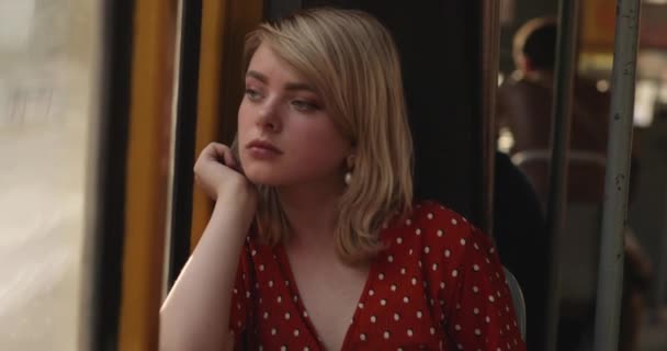 Tramvayda otururken pencereden dışarı bakan düşünceli genç bir kadının portresi. Kırmızı bluzlu, muhteşem altın sarısı saçları olan hoş bir bayan. 4k video — Stok video