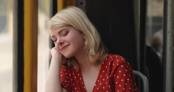 Portrait of young happy woman looking out a window, while she is sitting in the tram. Charming lady with a magnificent golden hair in red blouse thinking about something while she is riding in the — Stock Video