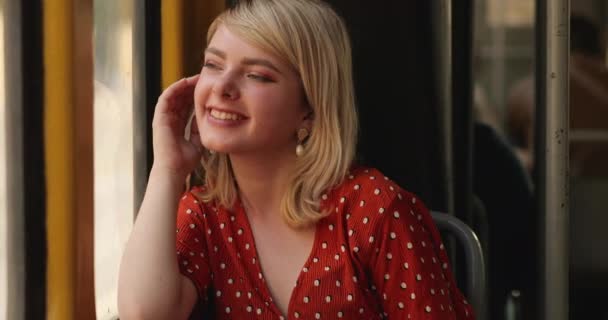 Portrait of young happy woman looking out a window, while she is sitting in the tram. Charming lady with a magnificent golden hair in red blouse thinking about something while she is riding in the — Stock Video