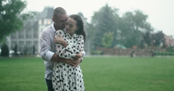 Vášeň v dešti. mladý milující pár objímající se v silném dešti. za nimi je zelené pole a velká budova. — Stock video