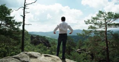 Bir adam kayanın kenarında duruyor. Dikiz aynası. Beyaz gömlekli ve kot pantolonlu bir adam güzel bir arka planda bir kayanın üzerinde duruyor ve ellerini kaldırıyor..