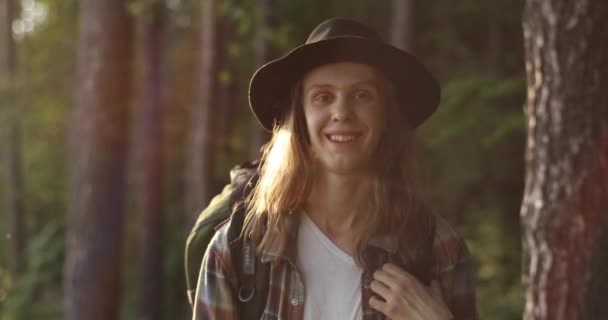 Портрет красивого молодого человека на шляпе, смотрящего в камеру и улыбающегося на закате в лесу. В поход. Саммер. 4К видео. Поездки . — стоковое видео