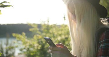 Büyüleyici, altın sarısı saçları, siyah güneş gözlükleriyle çekici genç bir bayan doğayla iç içe, akıllı telefonuyla bir şeyler yapıyor. Kamp, seyahat, doğa, orman. 4K video