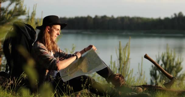 Beau jeune homme sur un chapeau regardant la carte, pensant à son itinéraire à travers la forêt. Camping. L'été. Vidéo 4K . — Video