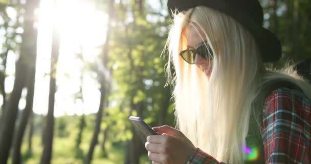 멋진 황금빛 머리에 검은 선글라스를 낀 젊은 여성을 매력적 인 아가씨가 자연을 좋아하고 스마트폰으로 뭔가를하고 있습니다. 캠핑, 여행, 자연, 숲. 4K 비디오 — 비디오